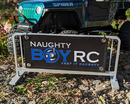 Naughty Boy RC Scale Mini Banner 3.5"x8" displayed in front of a radio control off-road vehicle, showcasing a sturdy stand and clear branding for rc car enthusiasts.
